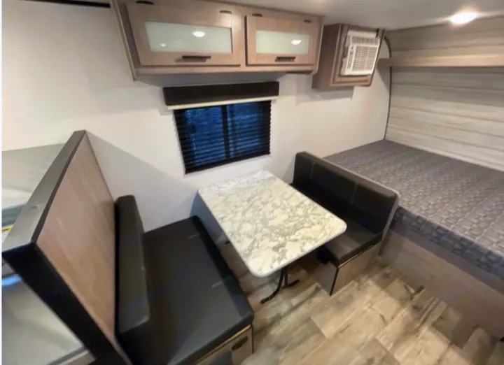 Compact RV rental interior featuring a dinette table, comfortable bed, overhead cabinets, and AC unit for cozy travels.. Coleman 17B 2023