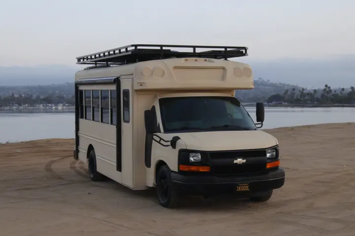 This tan bus-style RV rental with a roof rack is parked on a sandy shore by a calm bay, ready for adventure.. Chevrolet Express 2011