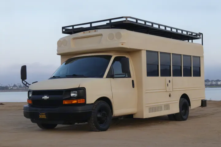 This tan, custom bus RV is parked on sand near water. An adventure-ready vehicle perfect for an rv rental.. Chevrolet Express 2011