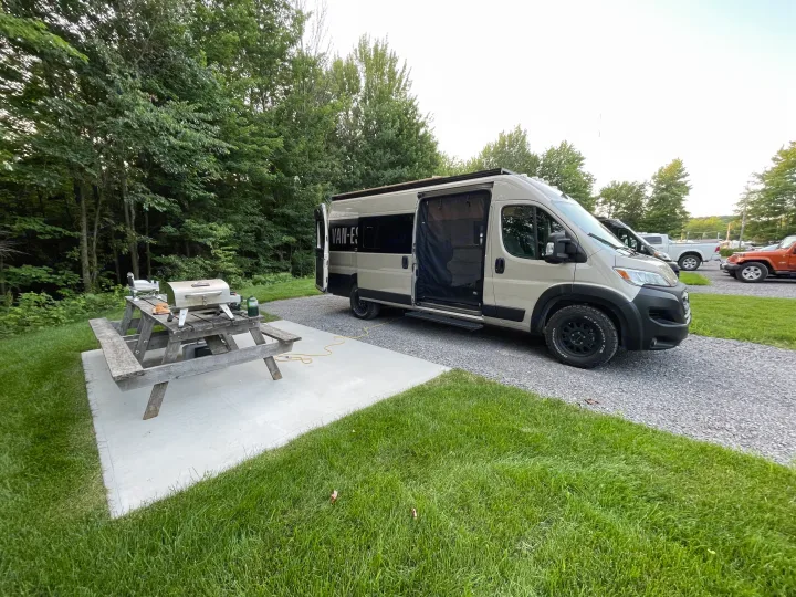 Peaceful RV rental campsite: A beige camper van, picnic table with grill on concrete pad, surrounded by grass & trees.. Ram Promaster 3500 2023