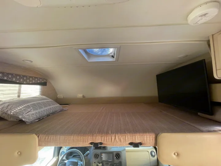 RV rental interior showing the cab-over bunk bed with a pillow, TV, and ceiling vent. View towards the driver's cabin.. Thor Freedom Elite 2022