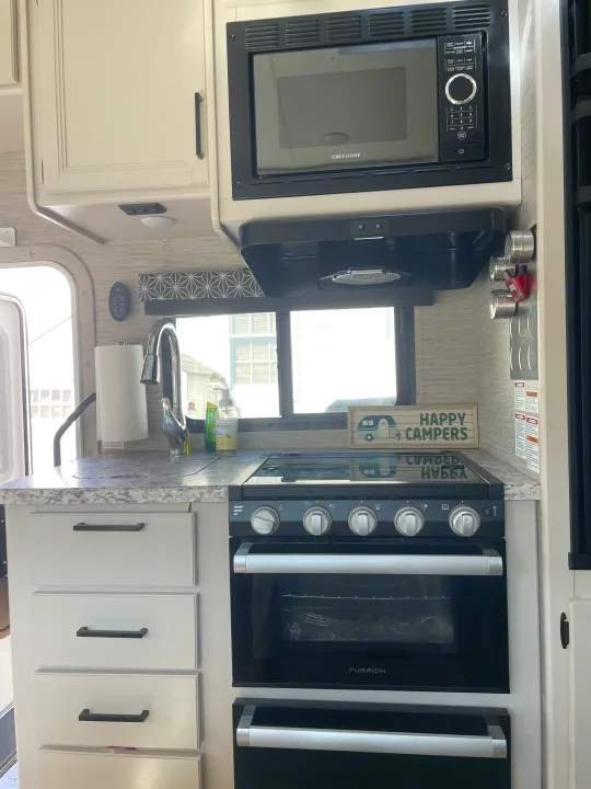 Clean RV rental kitchen: Greystone microwave, Furrion oven, sink, and "Happy Campers" sign. Ready for travel.. Thor Freedom Elite 2022