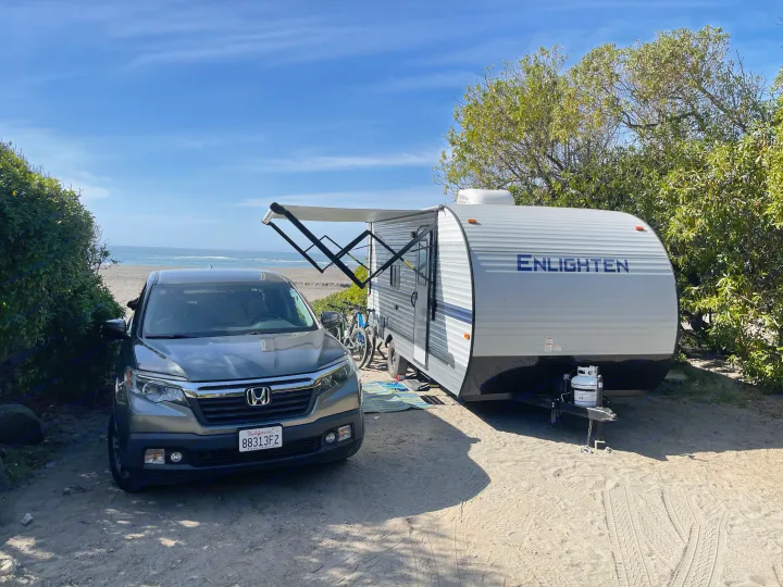 Coastal camping scene: A grey car and 'Enlighten' RV with awning extended, parked on sand by the beach. Consider an RV rental!. Gulf Stream Enlighten 2021