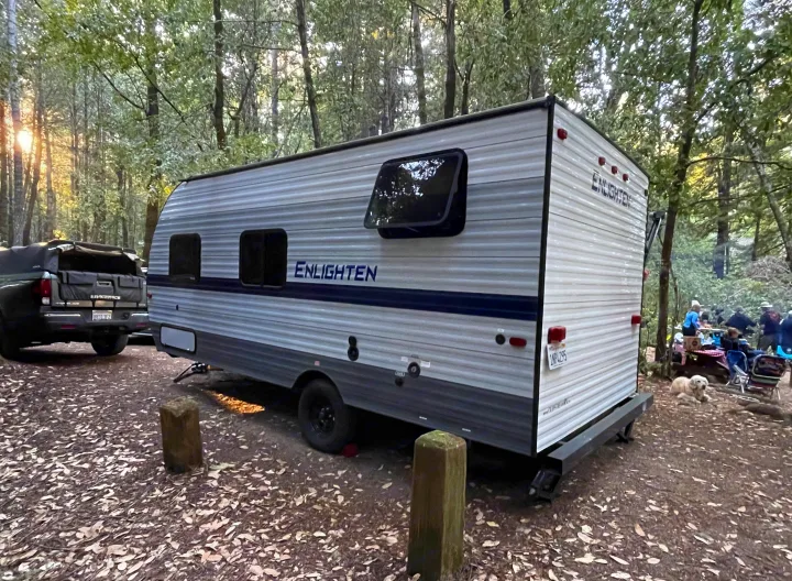 An Enlighten RV rental travel trailer is nestled in a forest campground with a truck, a dog, and people enjoying the outdoors.. Gulf Stream Enlighten 2021