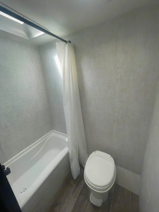 Clean RV rental bathroom with white bathtub, toilet, and shower curtain. Features light grey textured walls and wood-look flooring.. Gulf Stream Enlighten 2021