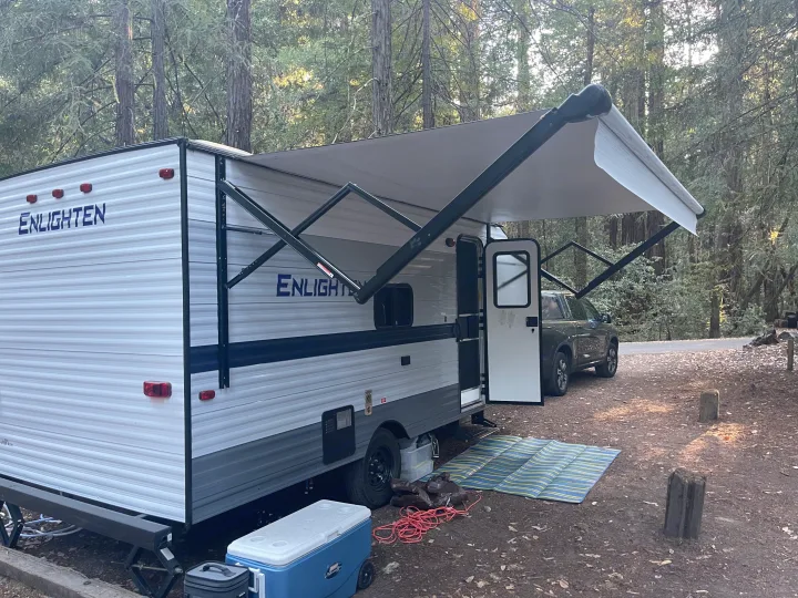 RV rental set up in a wooded campsite with awning extended, showing a cool blue cooler and truck nearby.. Gulf Stream Enlighten 2021