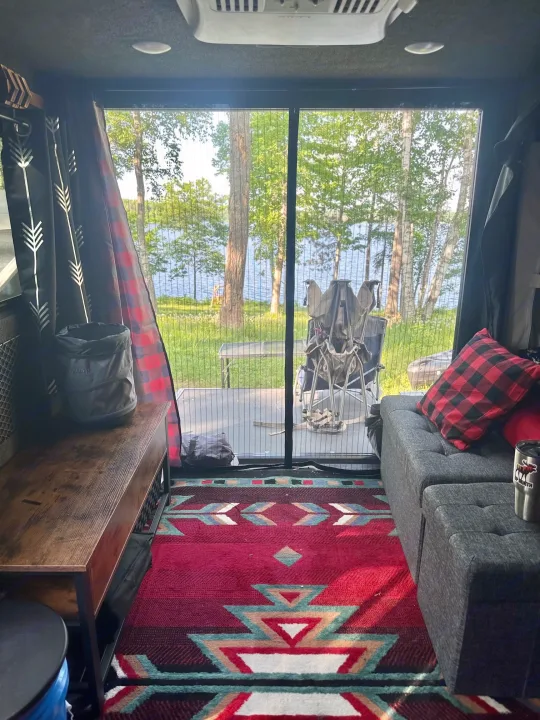 Inside an RV rental: a cozy space with a colorful rug and seating, looking out to a clear lake, green trees, and camping gear.. Intech Explore 2023