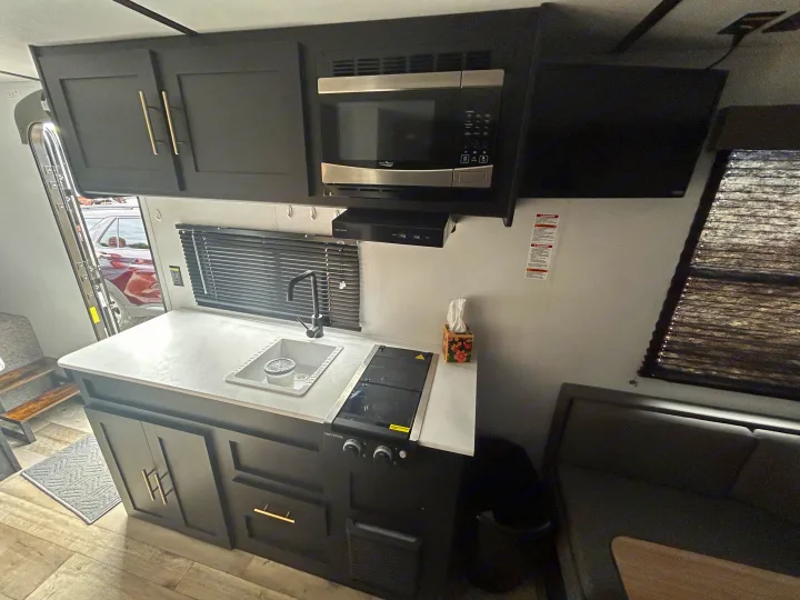 RV rental kitchen: modern dark cabinetry, microwave, sink, cooktop, and TV. A compact and clean travel space.. Keystone RV Springdale 2023