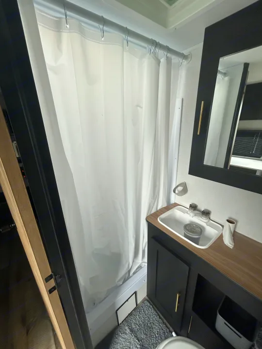 This RV rental bathroom features a white shower curtain, dark vanity with a sink, and a black-framed mirror.. Keystone RV Springdale 2023