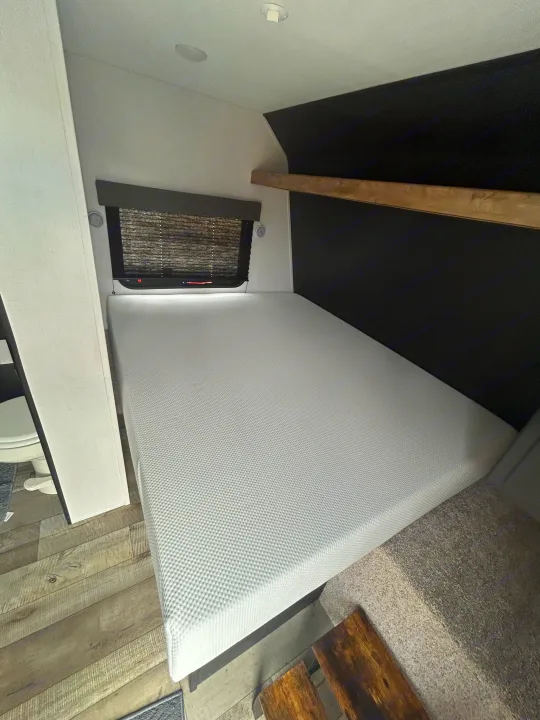RV rental bedroom: A bright, compact space featuring a fresh white mattress, window, and stylish wooden shelf.. Keystone RV Springdale 2023