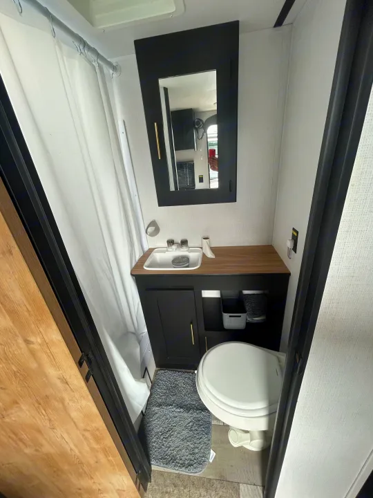 Inside an RV rental bathroom: white shower curtain, black vanity with sink, mirror, and toilet. Small, clean space.. Keystone RV Springdale 2023
