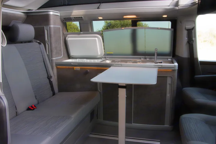 Spacious RV rental interior featuring a compact kitchenette with fridge, stove, and sink, plus comfortable seating and a dining table.. Volkswagen California Ocean 2025