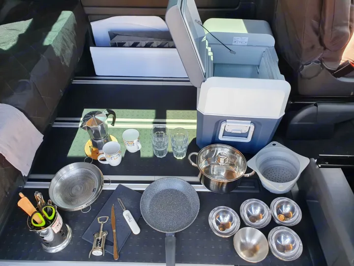 RV rental's mobile kitchen: a cooler, Moka pot, pot, pans, cutlery, and bowls laid out in a spacious vehicle interior.. Volkswagen Transporter California Beach 2019