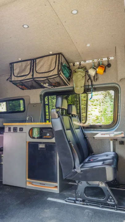 Inside an rv rental: custom camper van interior with a kitchen, refrigerator, flexible seating, and overhead storage. View of forest.. Mercedes-Benz Sprinter 2024