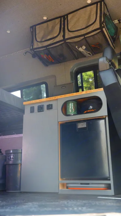 View inside an RV rental showing organized overhead storage, a built-in fridge, and countertop amenities.. Mercedes-Benz Sprinter 2024