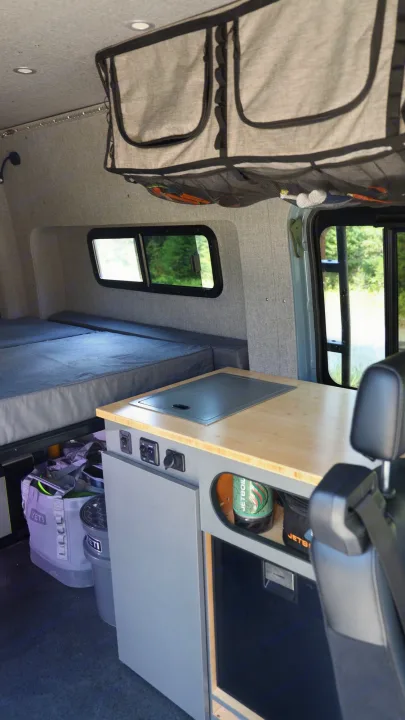 Modern RV rental interior showing a gray bed, light wood kitchen with sink, and practical overhead storage for adventure travel.. Mercedes-Benz Sprinter 2024