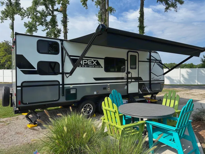 Relaxing RV rental setup: A white Apex travel trailer with an extended awning, colorful Adirondack chairs, and a patio table.. Coachmen Apex Nano 2024