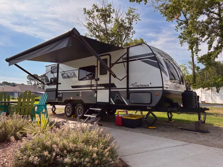This Coachmen Apex Nano-Off Grid RV rental with an extended awning is set up at a sunny campsite, chairs nearby.. Coachmen Apex Nano 2024