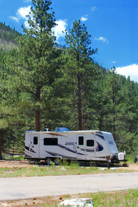 RV rental parked at a serene forest campsite, surrounded by tall pine trees and a mountain backdrop under a clear blue sky.. Lance Manufacturing Lance Manufacturing Trailer 2015