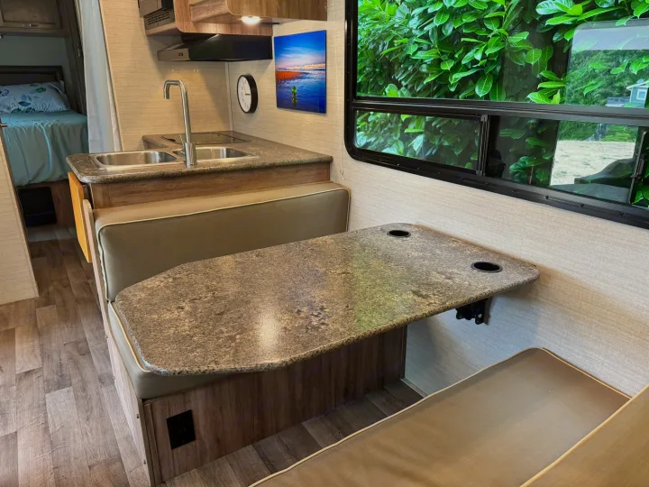 Modern rv rental interior: dinette with cup holders, kitchen with double sinks, and a bedroom beyond. Green scenery seen through window.. Jayco Redhawk SE 2019