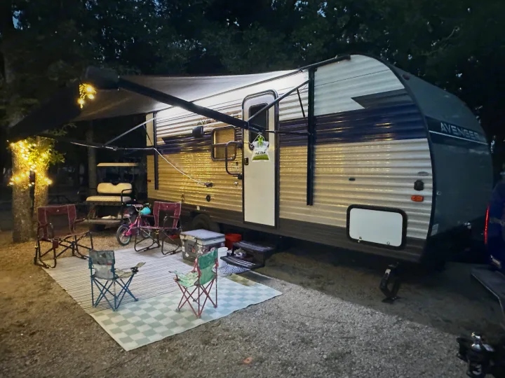 Cozy RV rental setup at a nighttime campsite, featuring illuminated awning, chairs, and mats. Great for family trips.. Prime Time Avenger - 17BHS 2022