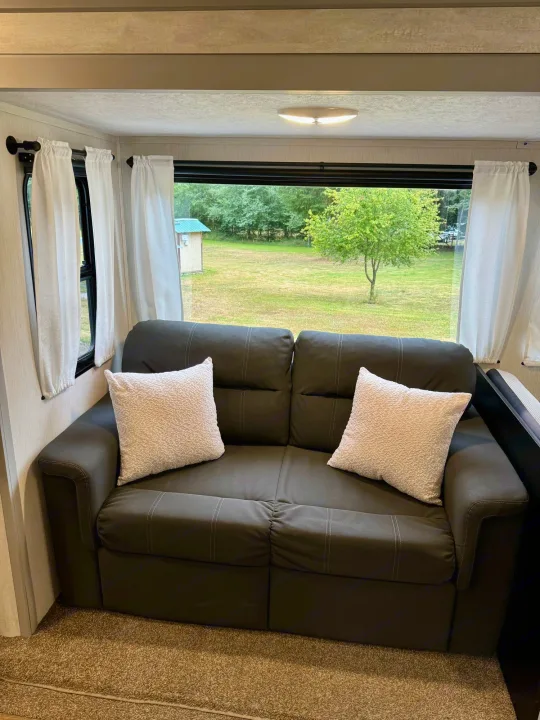 RV rental interior featuring a cozy dark sofa, white pillows, and a large window showcasing a grassy outdoor view.. Forest River Vibe 2021