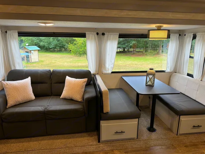 Bright RV rental interior showing a dark sofa, a dinette with storage, and large windows looking out to nature.. Forest River Vibe 2021