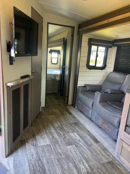 RV rental's inviting living area: gray recliners, TV, and wood flooring, with a glimpse into the connected bedroom.. Crossroads sunset trail superlite 2022