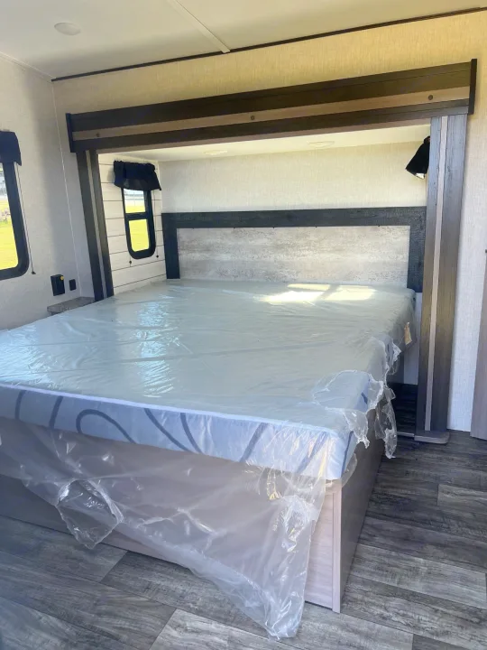 RV rental bedroom: Modern interior with a new mattress wrapped in plastic, wood-look headboard, and bedside shelf. Ready for travel.. Crossroads sunset trail superlite 2022