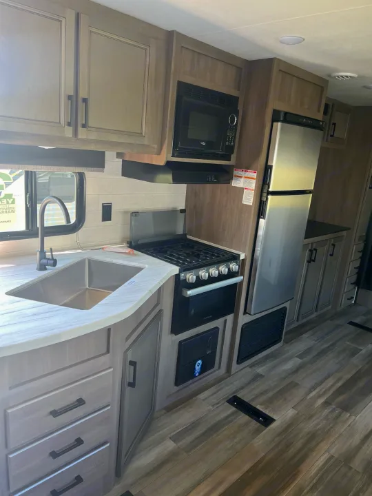 RV rental kitchen with a stainless steel sink, gas stove, microwave, and large refrigerator in modern light wood cabinets.. Starcraft Autumn Ridge 2023