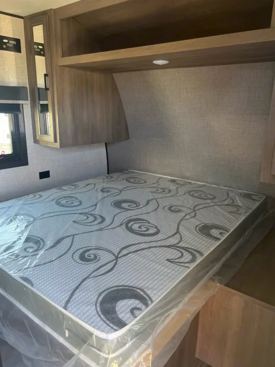 RV rental bedroom features a new mattress in plastic, overhead storage, and a window, ready for your travels.. Starcraft Autumn Ridge 2023