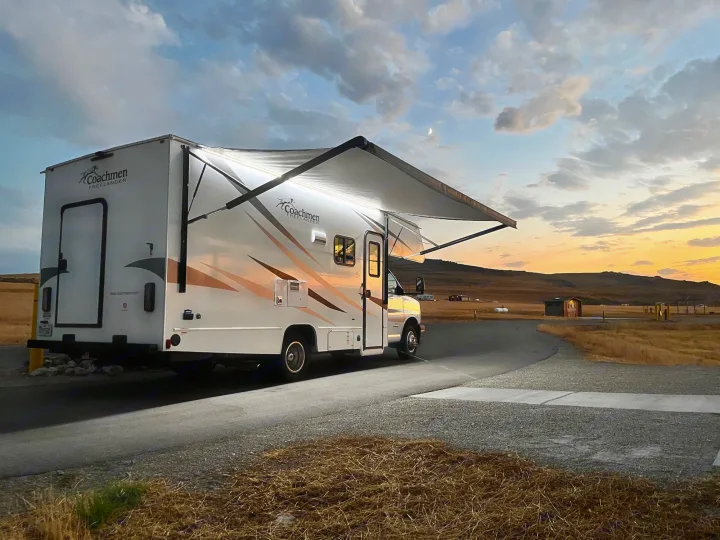 A white Coachmen rv rental at dusk, its lit awning bright against the sunset sky with a crescent moon overhead.. Coachmen Freelander 2020