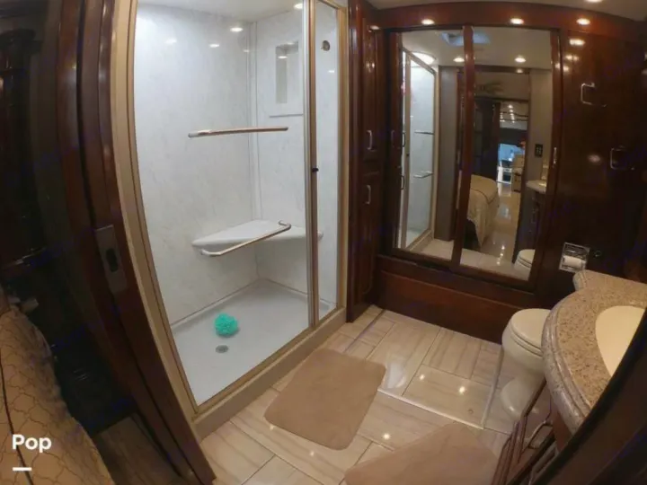 This RV rental bathroom has a glass shower, tiled floor, wood vanity, and toilet. Mirrors reflect a bed.. American Coach Revolution 2017