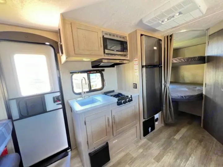 RV rental interior: A compact kitchen (sink, stove, microwave, fridge) and bunk beds are visible, with an entry door on the left.. Coachmen Catalina 2022