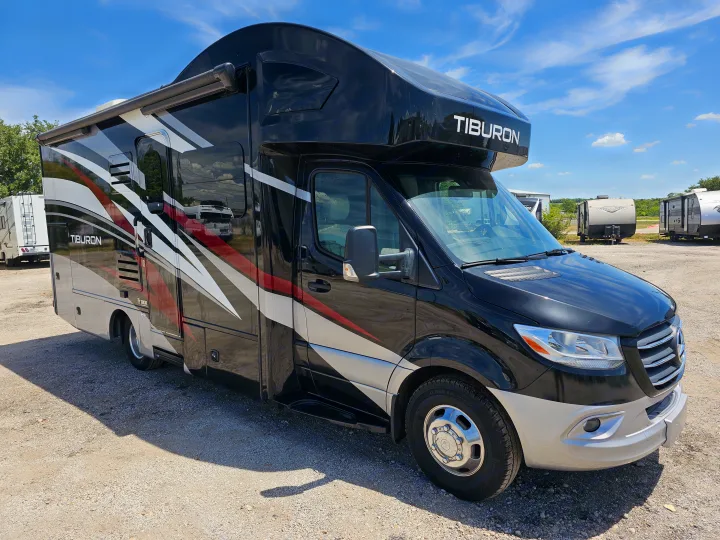 This black and silver Tiburon Class C RV rental, with an awning extended, is parked on a gravel lot on a sunny day.. Thor Motor Coach Other 2023