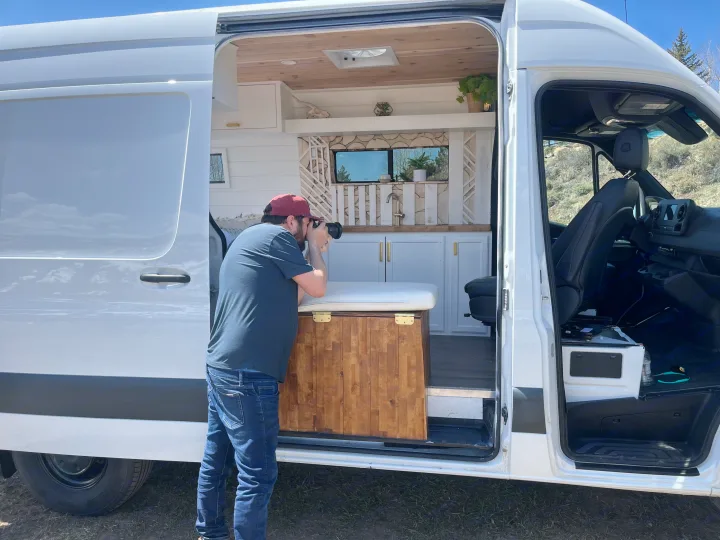Man photographs the detailed, custom white camper van interior with wood ceiling, cabinets & sink. Perfect for an rv rental.. Mercedes-Benz Sprinter-Loaded 2022
