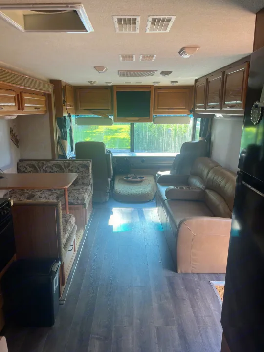 Spacious RV rental interior with a patterned dinette, wood floors, and tan leather sofa, leading to the bright front cabin.. Fleetwood Flair 31A 2005