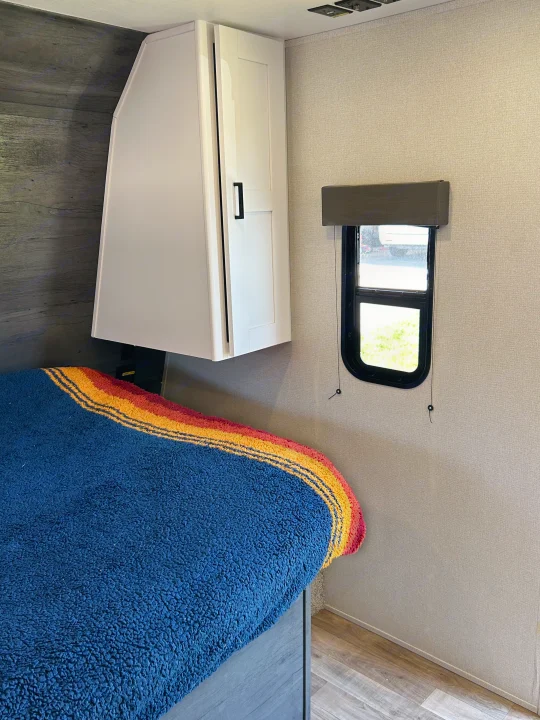 A cozy RV rental bedroom features a bed with a striped blanket, overhead storage, and a window.. Keystone RV Hideout 2022