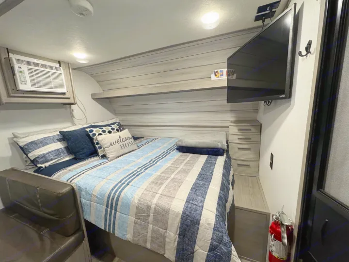 An RV rental bedroom setup: striped bed, mounted TV, AC unit, and a visible fire extinguisher for safety.. Coleman Lantern LT 17B 2024