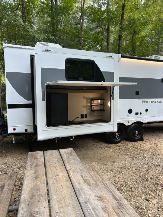 Wildwood RV rental: A travel trailer with an outdoor kitchen extended, parked by a picnic table at a wooded campsite.. Forest River RV Wildwood 31KQBTSX 2025