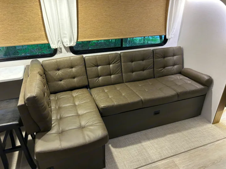 Comfortable L-shaped sectional sofa inside an RV rental. Features tufted cushions, under-seat storage, & scenic window views.. Forest River RV Wildwood 31KQBTSX 2025