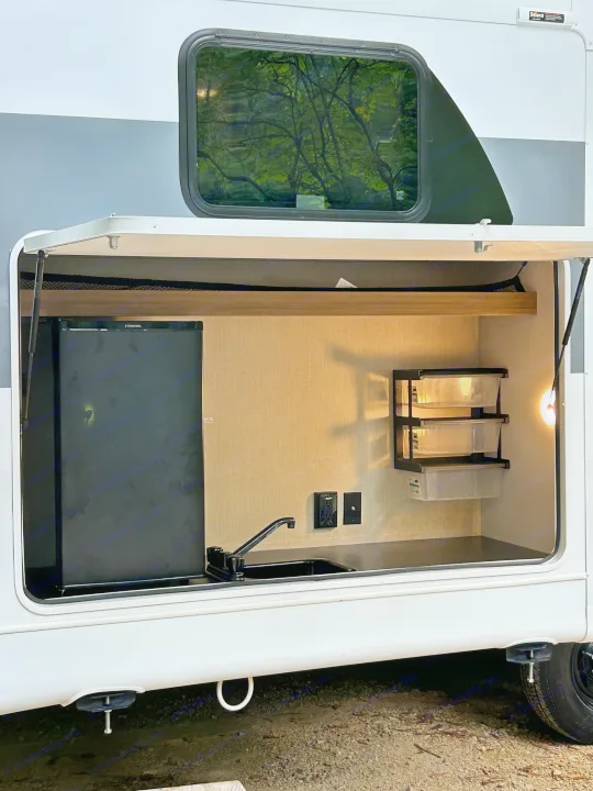 This rv rental's outdoor kitchen includes a fridge, sink, and tiered storage bins for easy camping.. Forest River RV Wildwood 31KQBTSX 2025