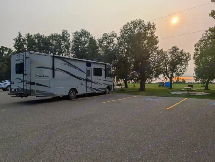 An RV rental parked in a paved lot by a lake, surrounded by trees. Hazy sky with sun, tents & picnic table.. Coachmen Pursuit 2021