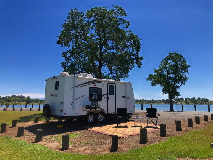 Scenic RV rental set up with outdoor seating by a lake, ideal for relaxing in nature under clear skies.. Forest River Rockwood Mini Lite 2012