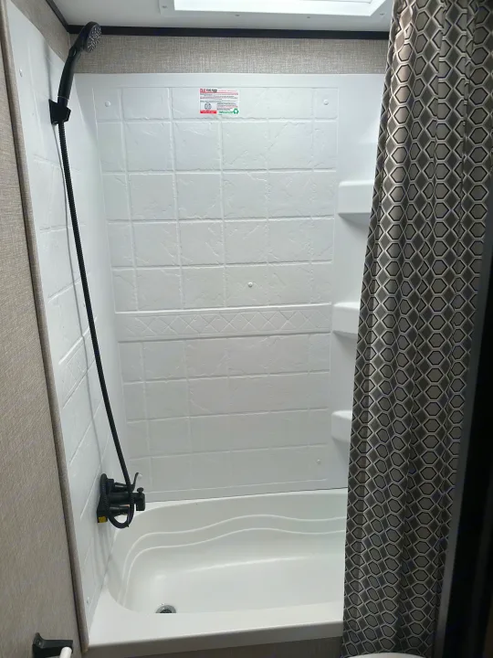 Inside an RV rental: a compact white shower and bathtub unit with a black handheld showerhead and geometric shower curtain.. Jayco Jay Flight 2022