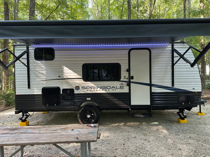 This rv rental is a white Springdale Keystone Mini travel trailer with blue awning lights, parked at a wooded campsite.. Keystone RV Springdale 2024