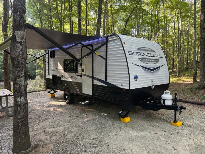 A white Springdale travel trailer, an **RV rental**, is parked at a scenic wooded campsite with its awning extended and lights on.. Keystone RV Springdale 2024