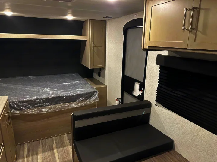 Inside a clean RV rental: a bed still wrapped in plastic, modern gray cabinets, and a small dinette area with black seating.. Keystone RV Springdale 2024