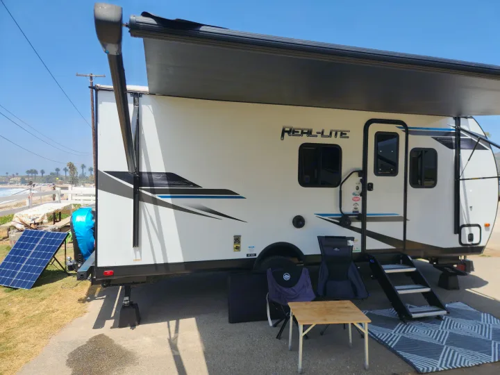 This RV rental features a Real-Lite travel trailer with an extended awning, solar panel, and outdoor setup by the sunny beach.. Palomino Real Lite Mini 2022