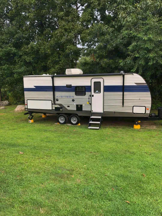 White & blue RV rental travel trailer parked on green grass with trees, stabilized for camping.. Forest River Prime Time Avenger 24BHS 2023